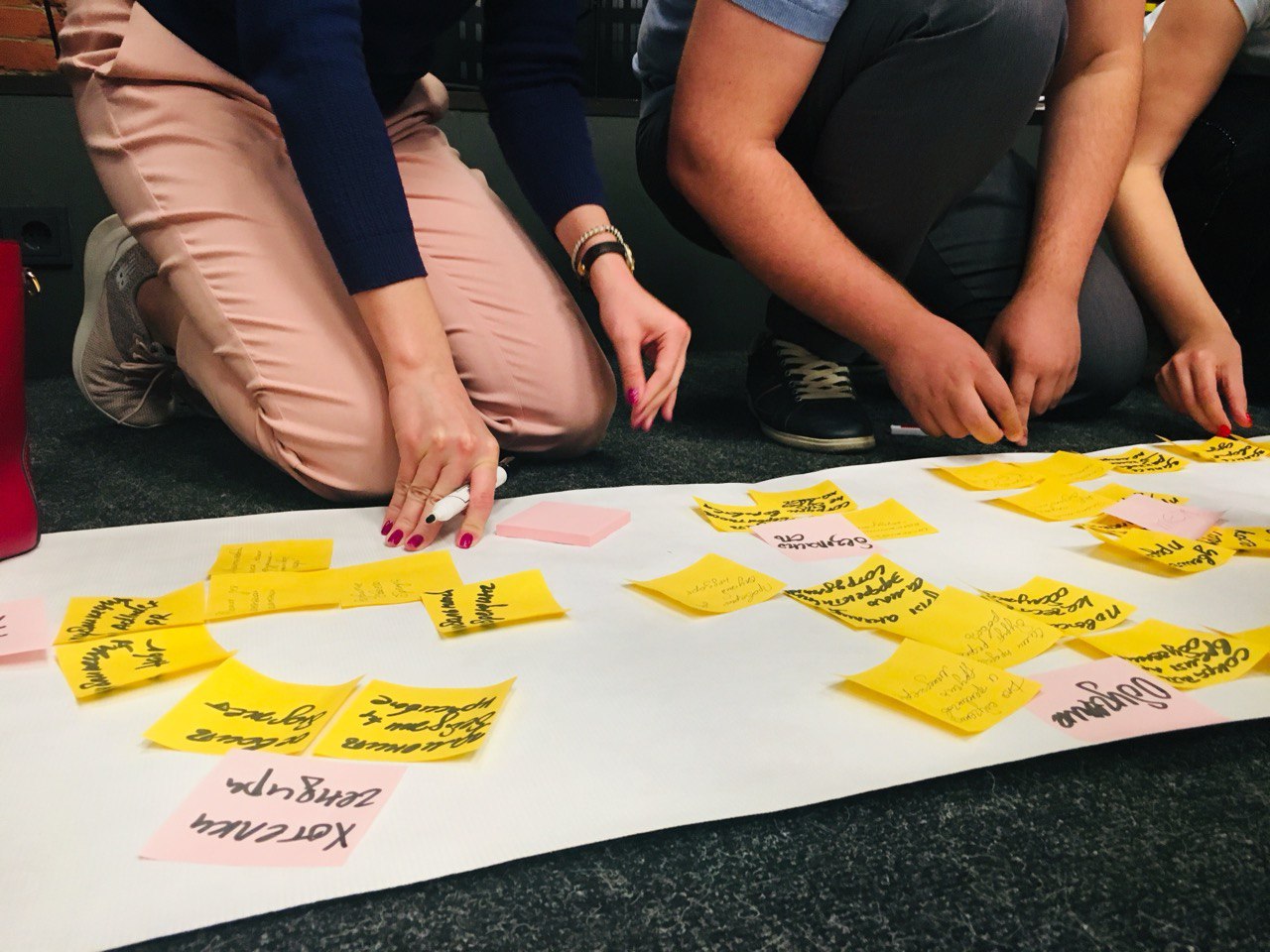 Несколько человек сидя на полу делают записи на стикерах, приклеенных на большом фрагменте рулонной бумаги